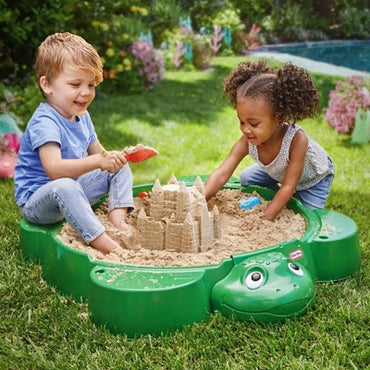 Two children playing with a sandcastle in a green sandbox shaped like a frog in a garden.